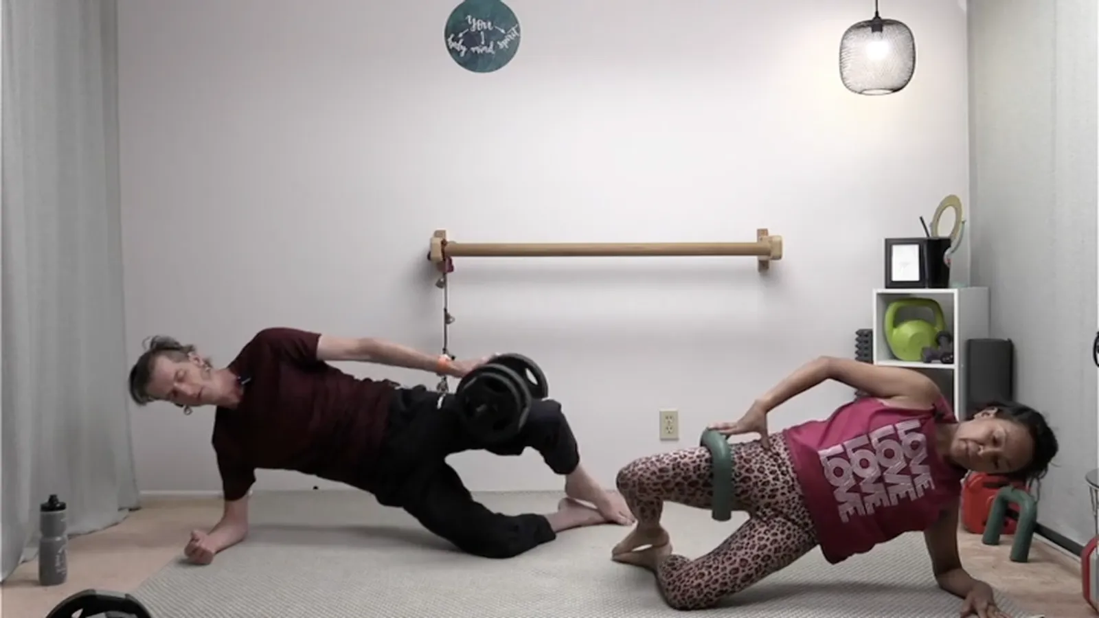 Two instructors holding side planks on mats in a home studio, one with a dumbbell and one with a weighted ring