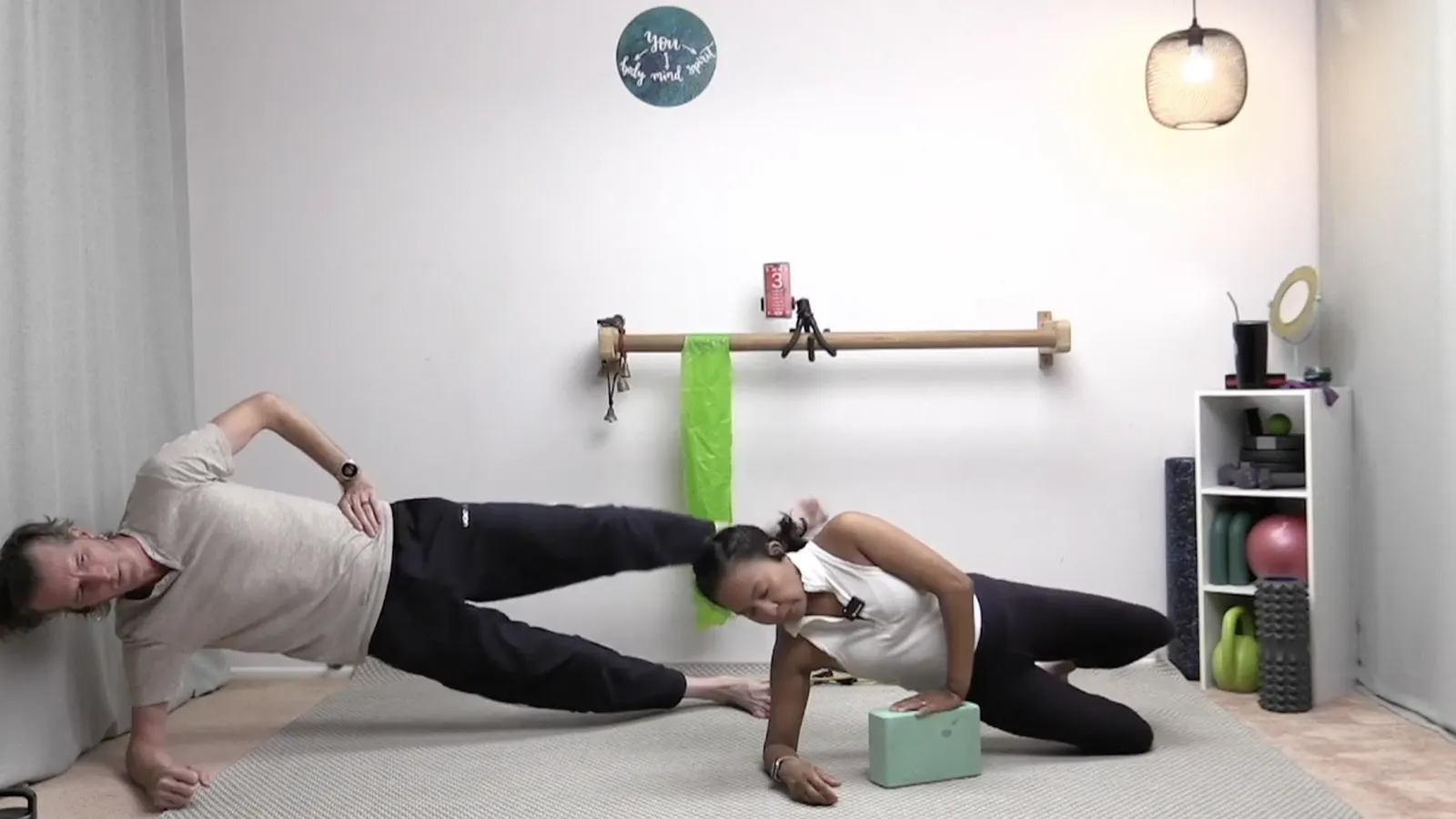 Two instructors doing side planks in a home studio, one supported by a foam yoga block