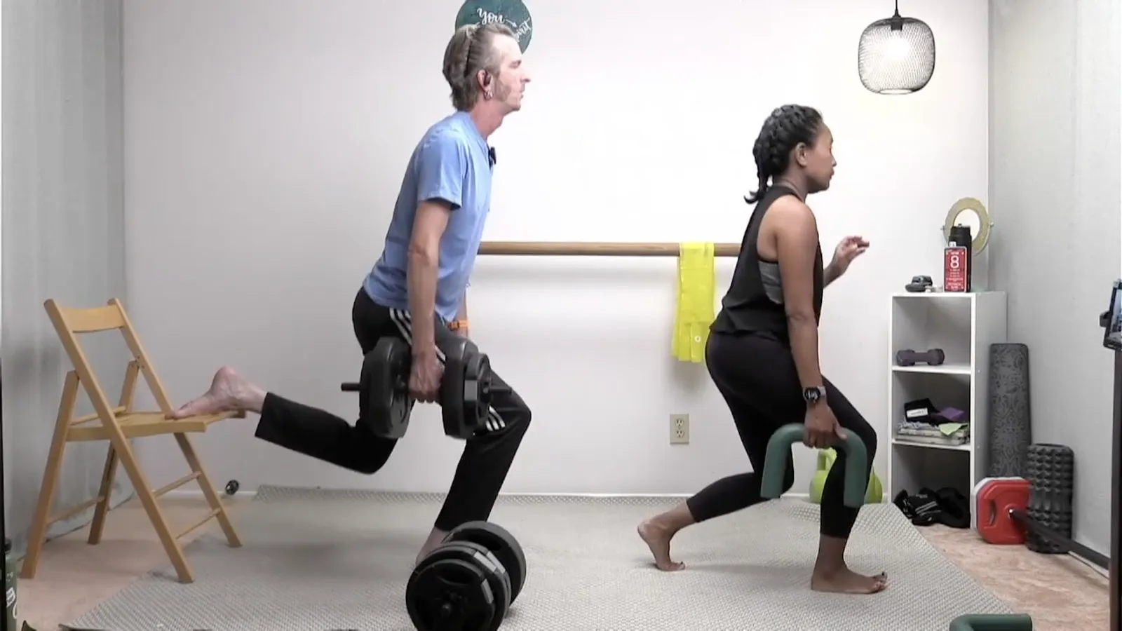 Two instructors doing split squats in a home studio, one with a rear foot on a chair holding dumbbells